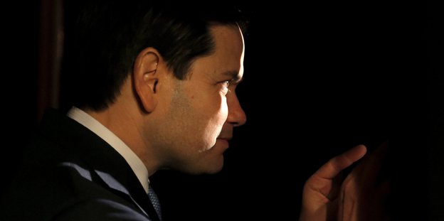 Marco Rubio steht im Dunkeln hinter einem Vorhang und blickt durch einen Spalt. Ein Lichtstreif fällt auf sein Gesicht.