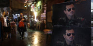 Nächtliche Szene, rechts im Vordergrund zwei Bilder des syrischen Machthabers Assad mit Sonnenbrille, dahinter Markt