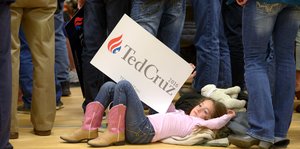 Ein Mädchen liegt auf dem Rücken auf dem Boden und hält ein Schild „Ted Cruz“ in die Höhe.