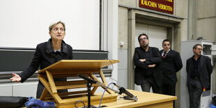 Judith Butler steht in einem Hörsal mit ausgebreiteten Armen hinter einen Lesepult.