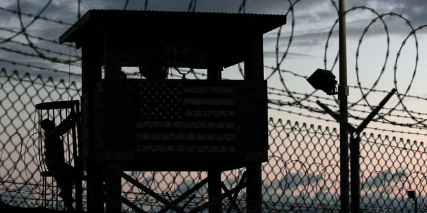 Die Silhouette eines Sicherheitszauns mit Natodraht vor dem Abendhimmel. Im Hintergrund klettert ein Soldat auf einen Wachtturm.