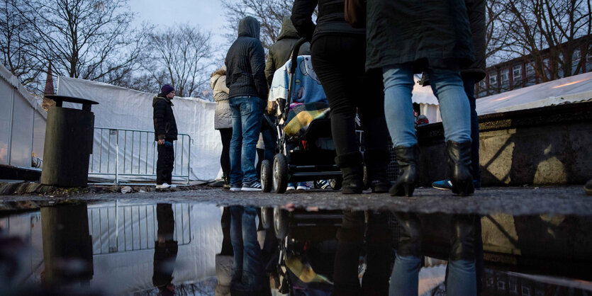 Menschen warten in einer Reihe vor einem Zelt. Neben ihnen ist eine Pfütze, in der sie sich spiegeln