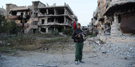 Mann mit Kalaschnikow und libyscher Flagge steht für zerstörten Häusern. In seinem Gesicht deutet sich ein Lächeln an.