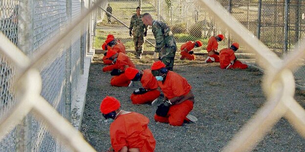 Etwa zehn Gefesselte Gefangene auf den Knien. Ihr Blick ist auf den Boden gerichtet. Zwischen ihnen laufen US-Militärs hin und her.
