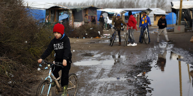 Ein Junge auf einem Fahrrad fährt durch Schlamm, dahinter das Flüchtlingslager in Calais