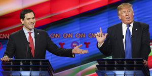 Marco Rubio und Donald Trump an zwei Rednerpodesten in einem Fernsehstudio. Rubio lächelt, Trump echauffiert sich mit gehobenem Zeigefinger.