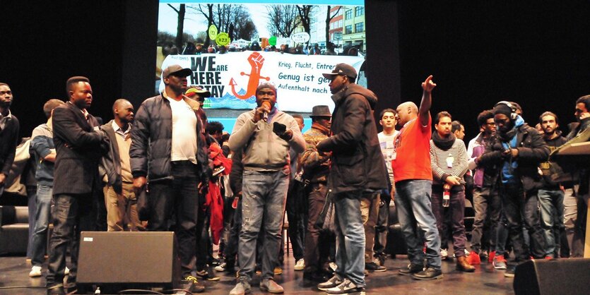 Flüchtlinge stehen auf einer Bühne der Theaterfabrik Kampnagel in Hamburg vor einem Plakat mit der Aufschrift "We are here to stay".