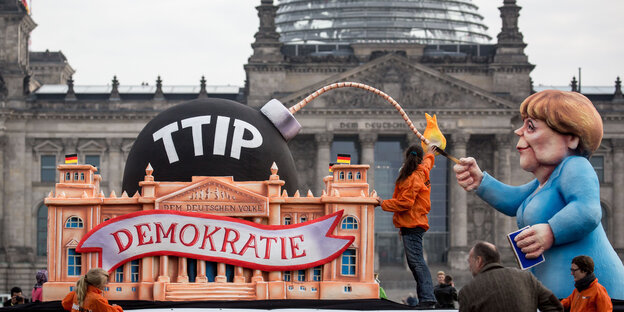 Anti TTIP Demo vor dem Bundestag. Eine Pappversion von Merkel zündet eine Pappbombe an, auf der TTIP steht