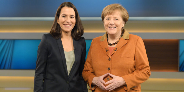 Anne Will steht lächelnd neben Angela Merkel, die mit ihren Händen eine Raute formt.