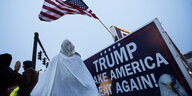 Ein Kritiker von Trump hat sich als Anhänger des Ku-Klux-Klans verkleidet und steht neben einem Schild „Trump make America great again!“.