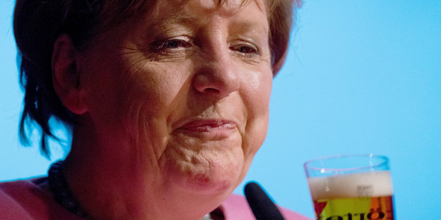 Merkel verzieht das Gesicht, vor sich ein Glas mit Flüssigkeit