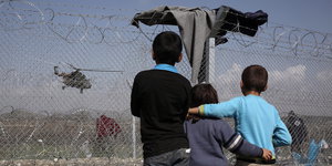 Drei Kinder, von hinten fotografiert, schauen durch einen Grenzzaun einem Helikopter zu