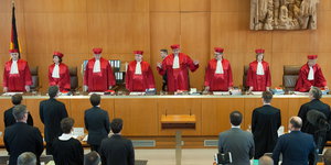 Die acht rotgekleideten Richterinnen und Richter stehen an einem langen Tisch vor dunkel gekleidetem Publikum
