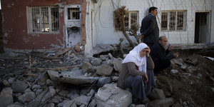 Eine Frau mit Kopftuch sitzt auf Trümmern vor zerstörten Häusern. Neben ihr sitzt ein Mann, dahinter steht ein zweiter Mann