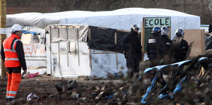 Bauarbeiter und Polizisten in Kampfmontur vor verbrannten Resten einer Hütte und einem Zelt, auf dem "Schule" steht