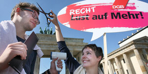 Eine Aktivistin und ein Aktivist halten ein Transparent auf dem "Recht auf Mehr" steht, eine Schere und eine großen Kamm vor dem Brandenburger Tor