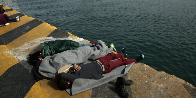 Menschen sind in Decken gewickelt. Sie liegen an einem Hafenbecken.