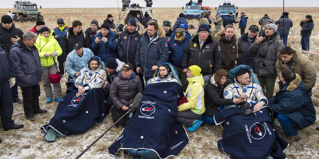 Drei Astronauten sitzen in Decken gehüllt vor einer Gruppe Menschen.