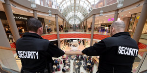 zwei Menschen in Schwarz mit Aufdruck "Security" auf dem Rücken blicken von einer Galerie runter in ein Einkaufszentrum