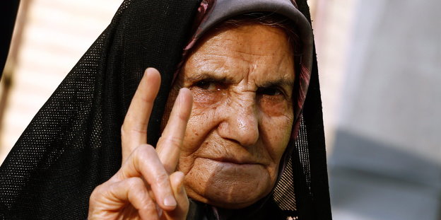 Eine alte Frau mit schwarzem Kopftuch hebt ihre Hand zum Victory-Zeichen.