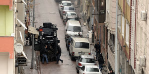 Straße mit parkenden Autos und einem gepanzerten Polizeifahrzeug in der Mitte, dahinter und in Hauseingängen bewaffnete Uniformierte