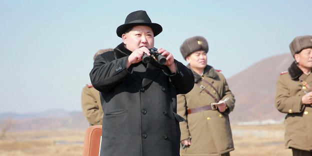 Kim Jong Un in dunklem Hut und Mantel, hält ein Fernglas in den Händen, hinter ihm stehen Soldaten in beigefarbenen Uniformen.