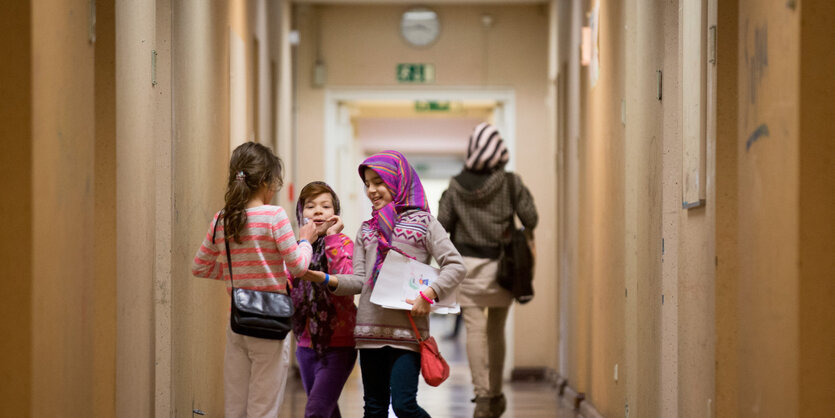 Drei lachende Kinder mit Kopftüchern und Täschchen in einem langen kargen Gang, dahinter eine Frau mit Kopftuch