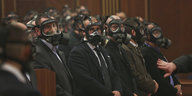 Männer in Anzügen und aufgesetzten Tränengasmasken sitzen in einer Reihe