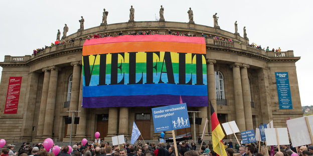 Gebäude mit Säulen, an dem ein riesiges Plakat in Regenbogenfarben mit der Aufschrift "Vielfalt" hängt, davor Menschen, einer davon mit einem Plakat auf dem steht: "Gender verschwendet Steuergelder"