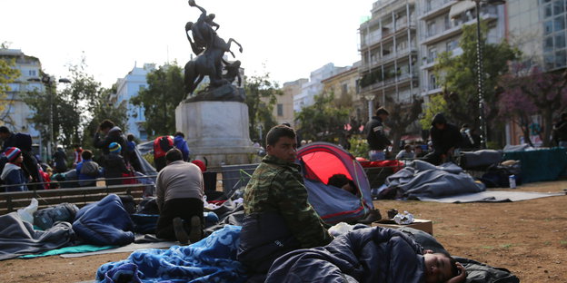 Menschen in Schlafsäcken auf der Erde vor einem Denkmal, dahinter Häuser
