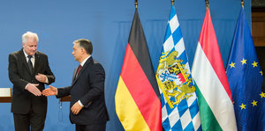 zwei Männer in Anzügen reichen sich die Hand, neben ihnen Fahnen von Deutschland, Bayern, Ungarn und der EU