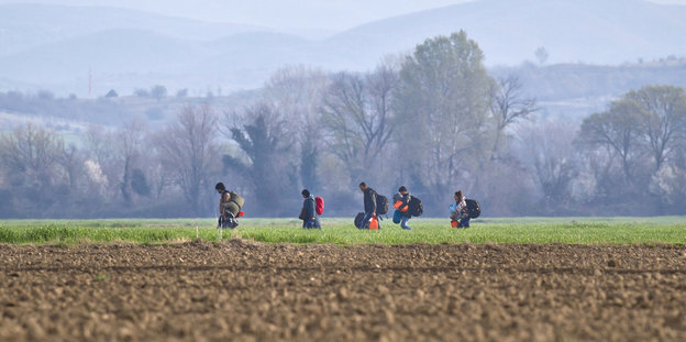 Flüchtlinge mit Taschen und Rucksack gehen durch eine Wiesen- und Ackerlandschaft. Straße rn