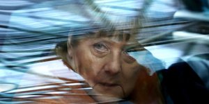 Frau Merkel blickt hinter einer reflektierenden Autofensterscheibe hervor.