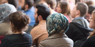 Frauen mit und ohne Kopftuch sitzen nebeneinander. Man sieht sie von hinten.