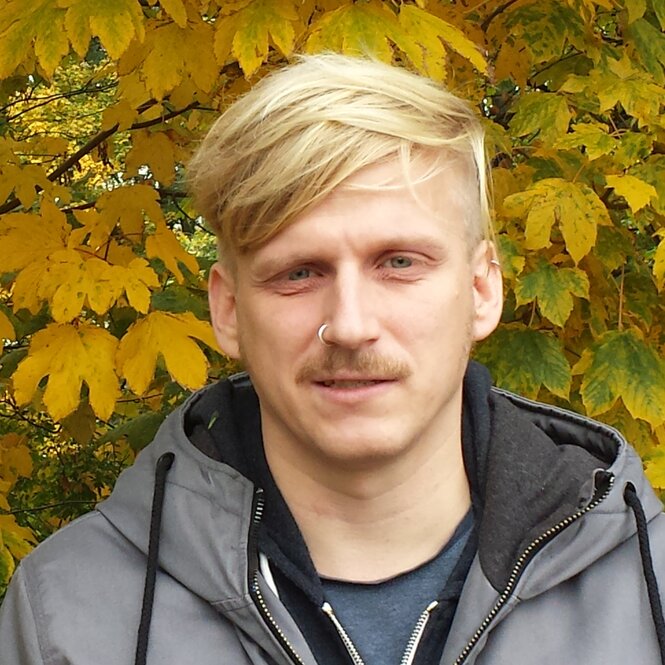 Ein Mann mit blonden Haaren steht vor einem Laubbaum mit gelben Blättern.