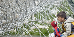 Ein Junge sitzt mit einer Spiderman-Puppe vor einem Stacheldrahtzaun.