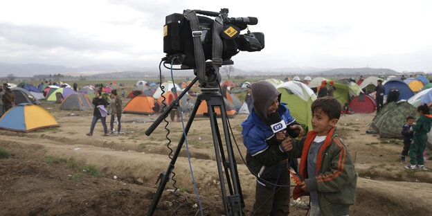 Zwei Kinder stehen neben einer Kamera auf einem Stativ, eines hält dem anderen ein Mikrofon entgegen.