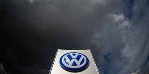 Die Sonne scheint auf das VW-Logo, am Himmel dunkle Wolken