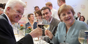 Winfried Kretschmann und Angela Merkel prosten sich mit einem Weinglas zu und schauen in die Kamera