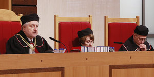 Drei Menschen in mit schwarzen Roben und Hüten sitzen hinter einem hohen Tisch
