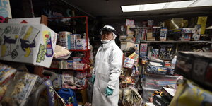 EIn MAnn mit Schutzkleidung steht in einem Laden zwischen Regalen mit unterschiedlichen Waren