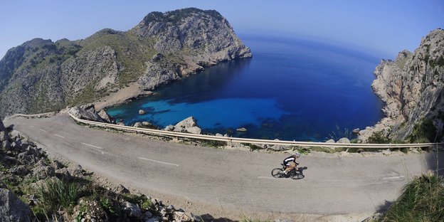 Eine Rennradfahrerin fährt auf einer Straße einen Berg hinauf. Hinter ihr liegen Berge, die Küste und das blaue Meer.