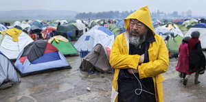 Künstler Ai Weiwei läuft in seinem gelben Regencape durch ein Flüchtlingslager mit verschienenen Zelten. Der Boden ist nass.