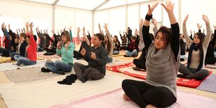 Frauen sitzen in einem Zelt im Yogasitz auf Matten und strecken die Arme nach oben.