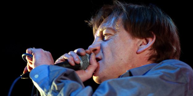 Der Sänger trägt ein blaues Hemd und hält ein Mikrofon in der Hand.