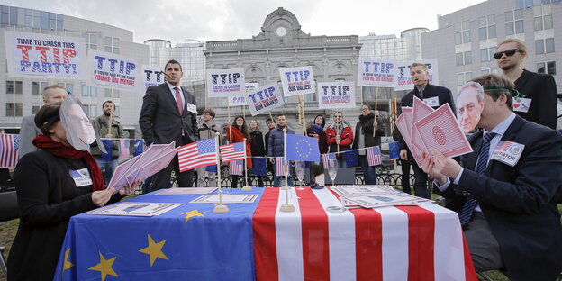 Auf einem Tisch liegt eine US-Fahne, daneben sitzen Aktivist_innen, die großeSpielkarten in der Hand halten, hinter ihnen stehen Aktivist_innen mit Anti-TTIP-Schildern.