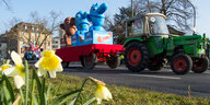 Ein Karnevalswagen stellt drei Figuren dar, die sich von blau nach braun verfärben. Er wird von einem grünen Traktor gezogen. Im Vordergrund stehen gelbe Blumen.
