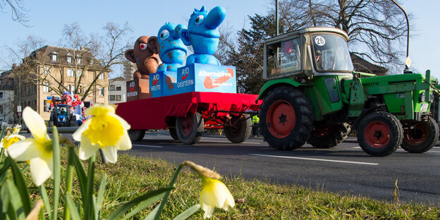 Ein Karnevalswagen stellt drei Figuren dar, die sich von blau nach braun verfärben. Er wird von einem grünen Traktor gezogen. Im Vordergrund stehen gelbe Blumen.