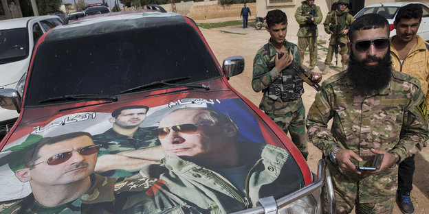 Uniformierte stehen neben einem Auto, auf dessen Motorhaube Putin und Assad abgebildet sind