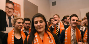 CDU-Anhänger_innen mit orangefarbenen Parteischals gucken betreten.